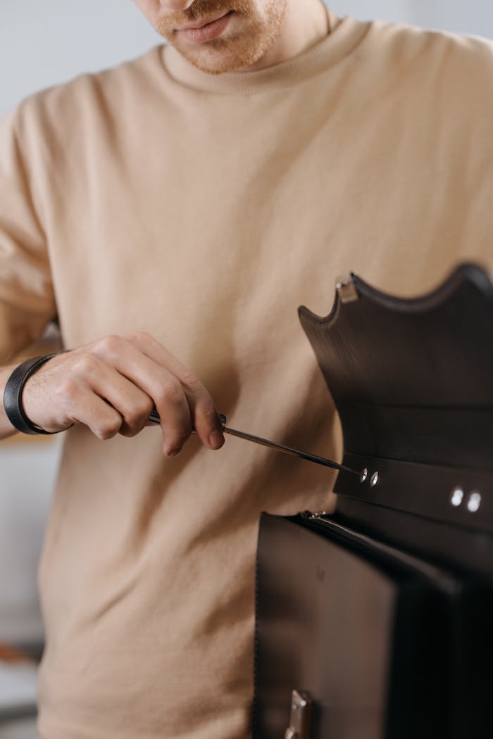 Close-up of a man using a screwdriver to repair a leather bag indoors.